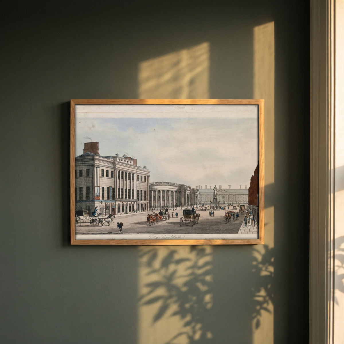 College Green, Dublin framed print in a natural oak frame on a dark green wall beside a window, dappled plant shadow