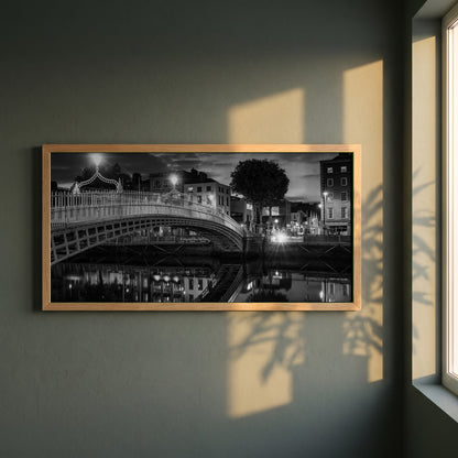 Night View of Ha'penny Bridge framed print in a natural oak frame on a dark green wall beside a window, dappled plant shadow