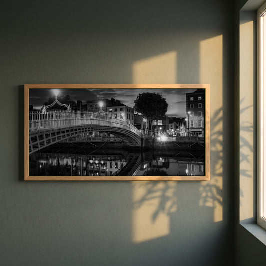 Night View of Ha'penny Bridge framed print in a natural oak frame on a dark green wall beside a window, dappled plant shadow