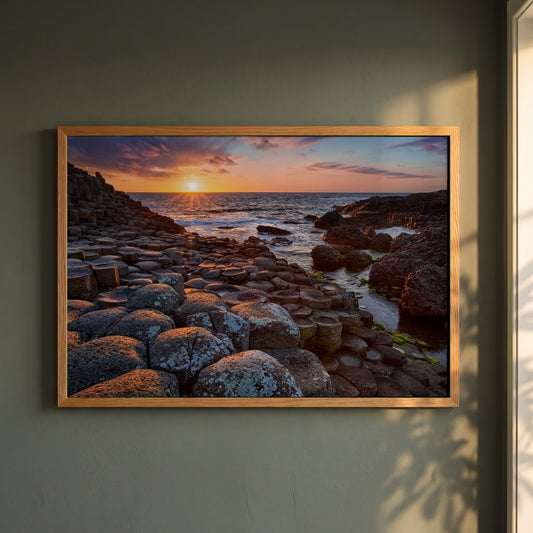 Giant's Causeway framed print in a natural oak frame on a dark green wall beside a window, dappled plant shadow
