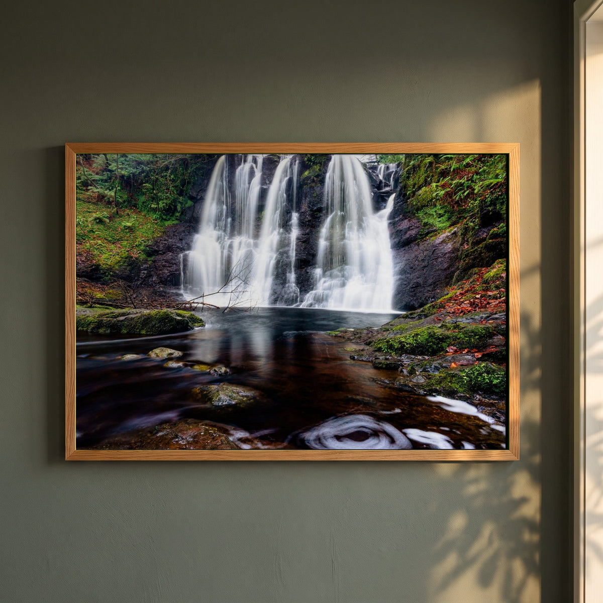 Glenariff Waterfall framed print in a natural oak frame on a dark green wall beside a window, dappled plant shadow