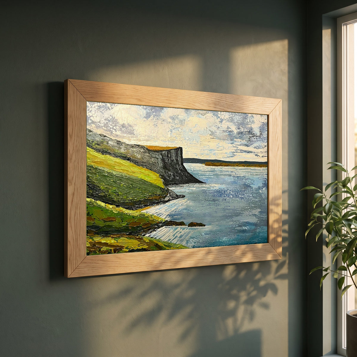 Murlough Bay & Fair Head framed print in a natural oak frame on a dark green wall beside a window, dappled plant shadow